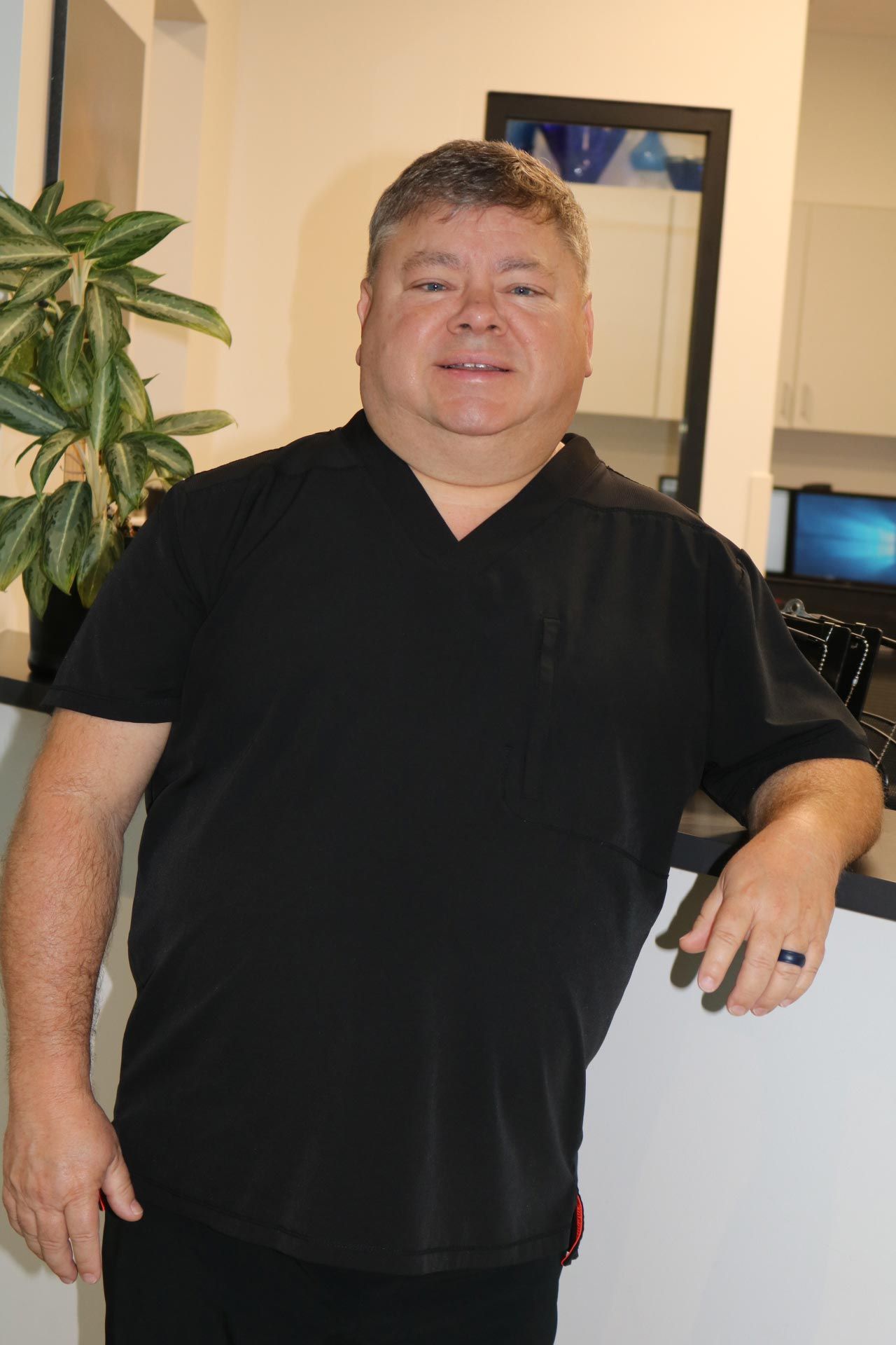 Man in black scrubs leans on a counter, smiling. Indoor setting with a plant and mirror.