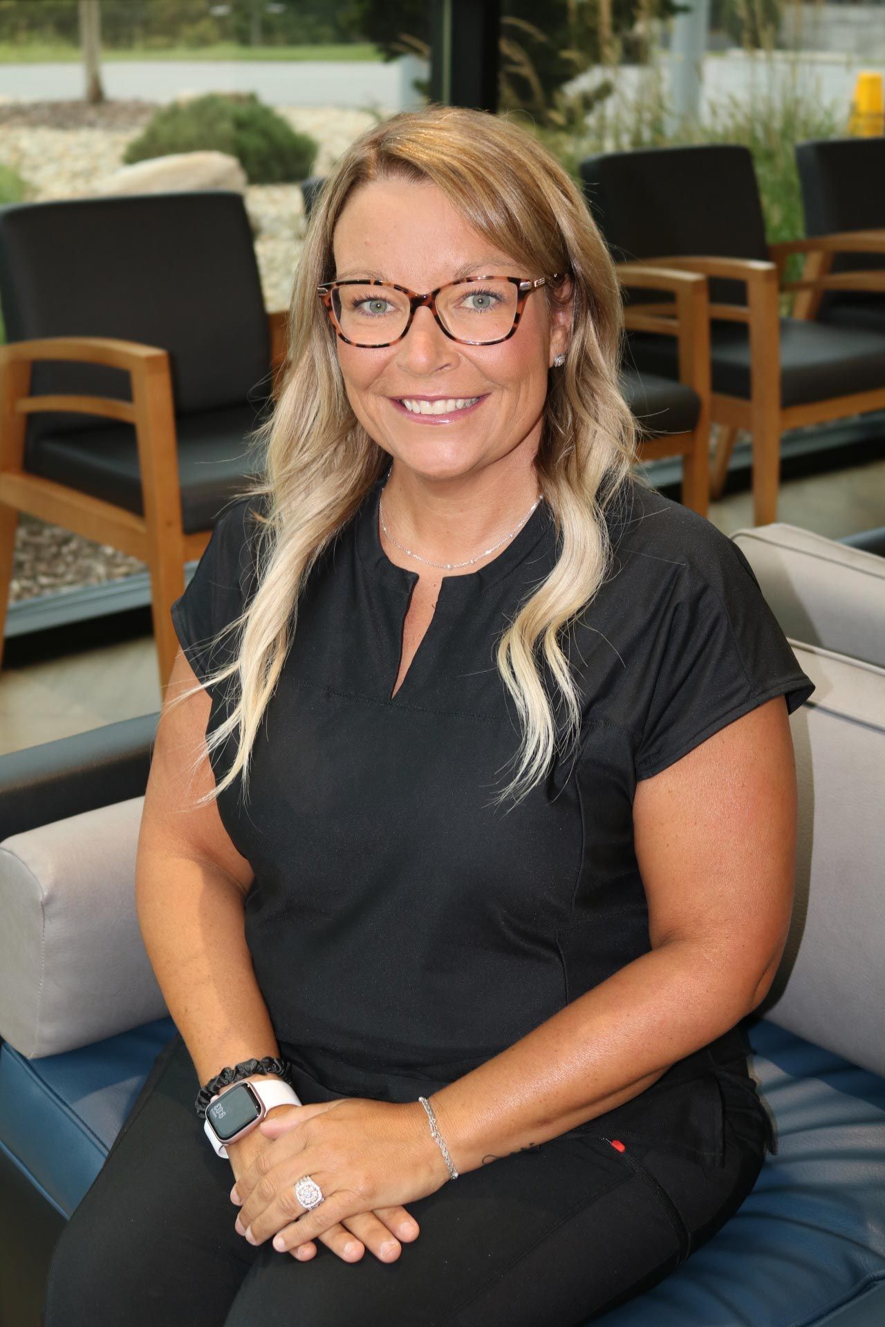 Woman with blonde hair wearing glasses, black top, seated, smiling.