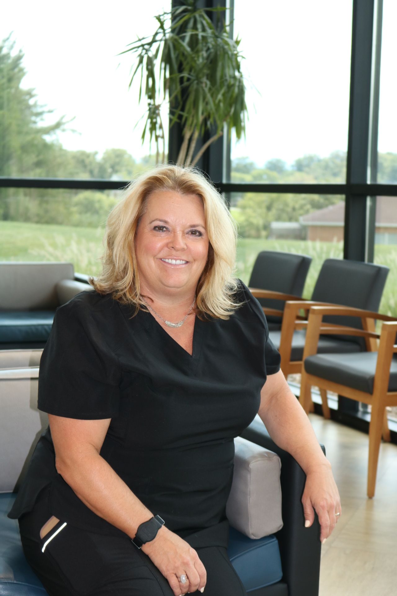 Woman in black scrubs smiling, seated in a waiting room with large windows and chairs.