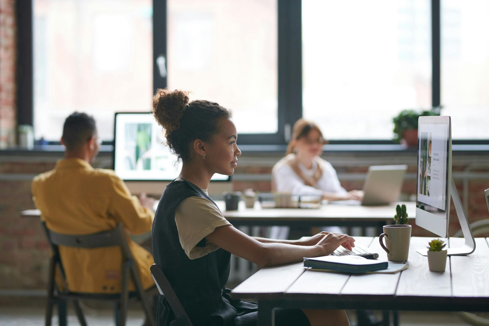 Woman working on a computer in an office, other people in background.