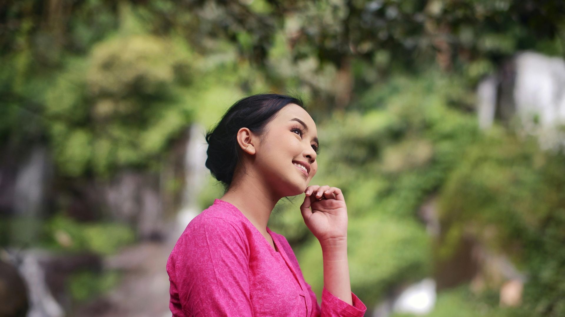 Woman in a bright pink top smiling outdoors in a green garden, hand near her face.