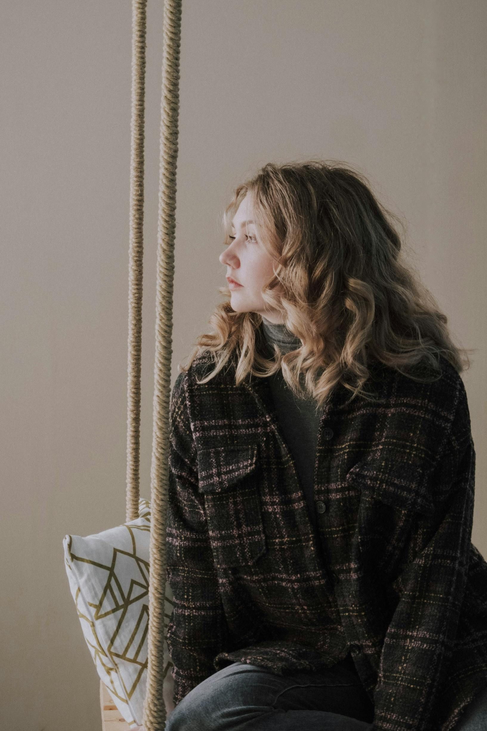 Woman in a dark plaid coat sitting by a window, gazing to the side, with a geometric cushion beside her