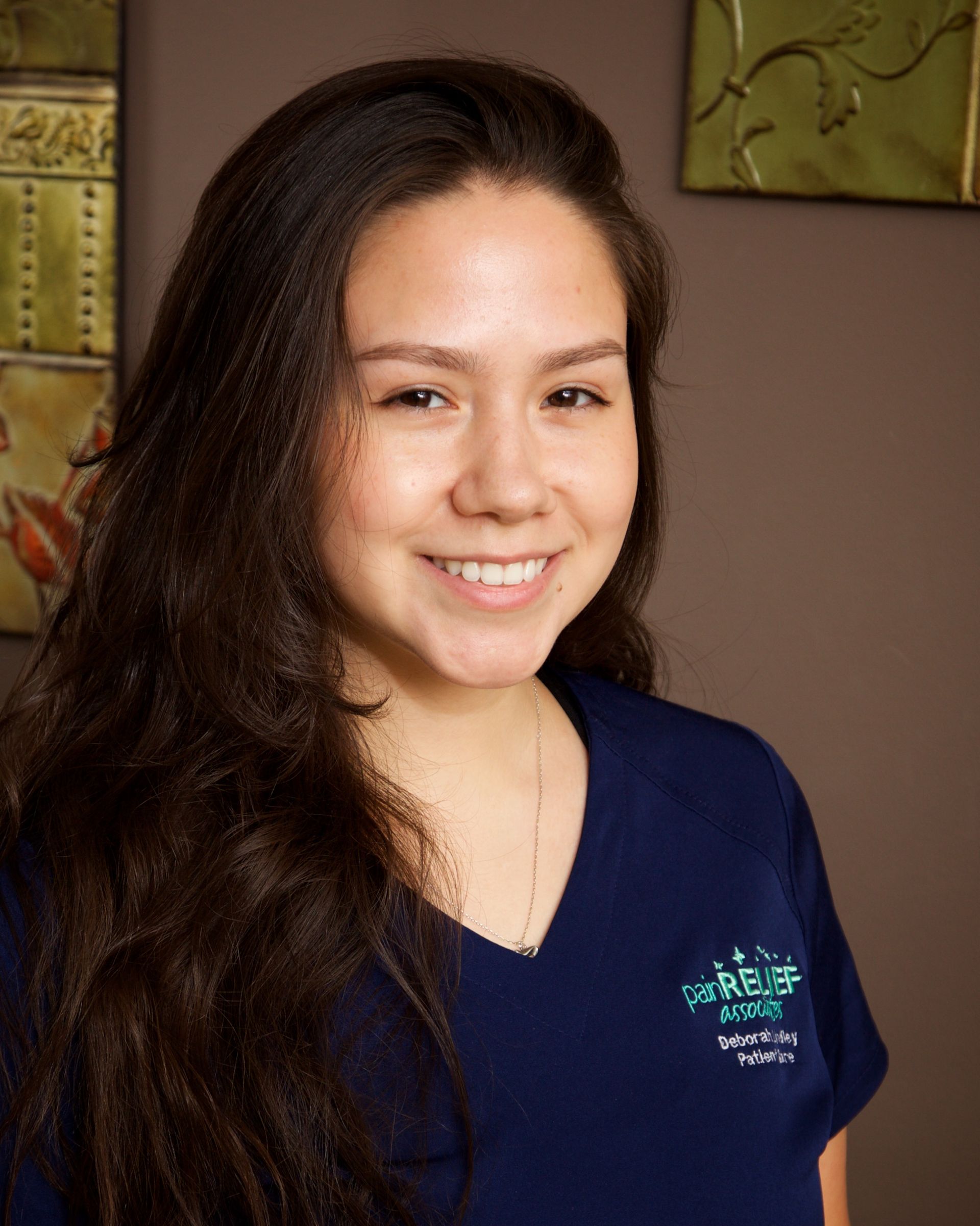 Woman with long dark hair smiles, wearing a blue scrub top, indoors.