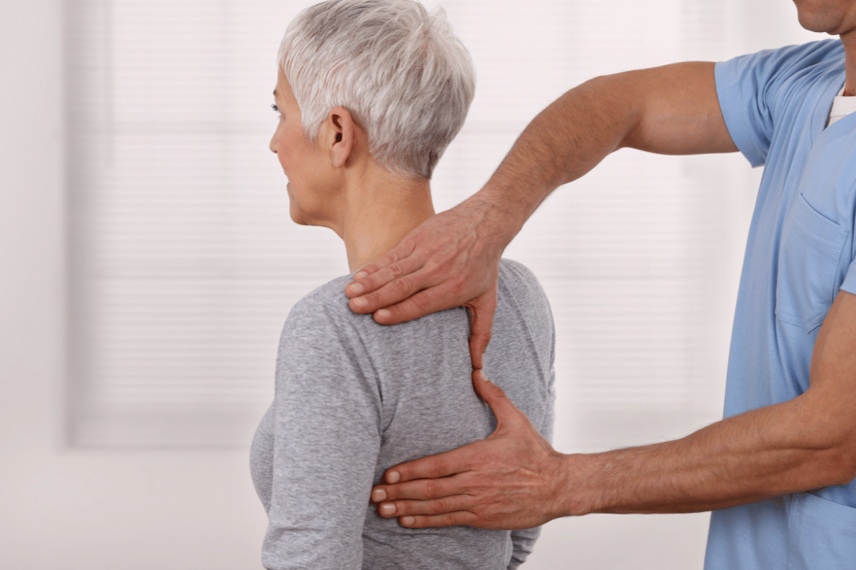 Physiotherapist examining older woman's back. They are indoors, wearing light-colored clothing.