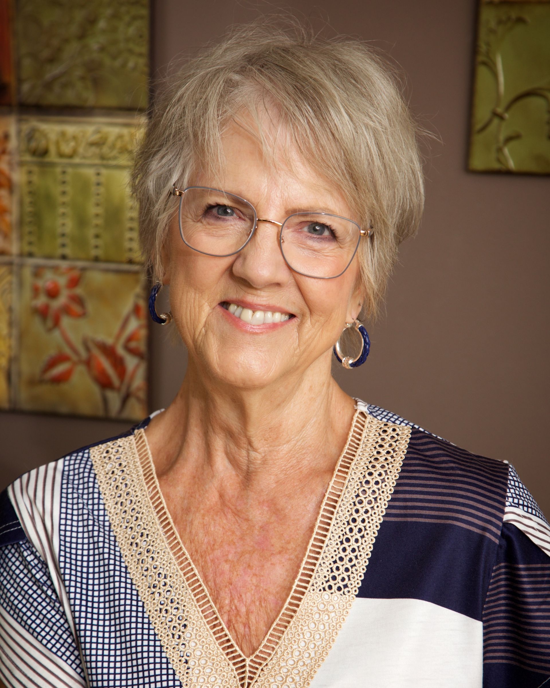 Woman with short gray hair, glasses, and smiling, wearing a patterned shirt, stands near a decorative wall.