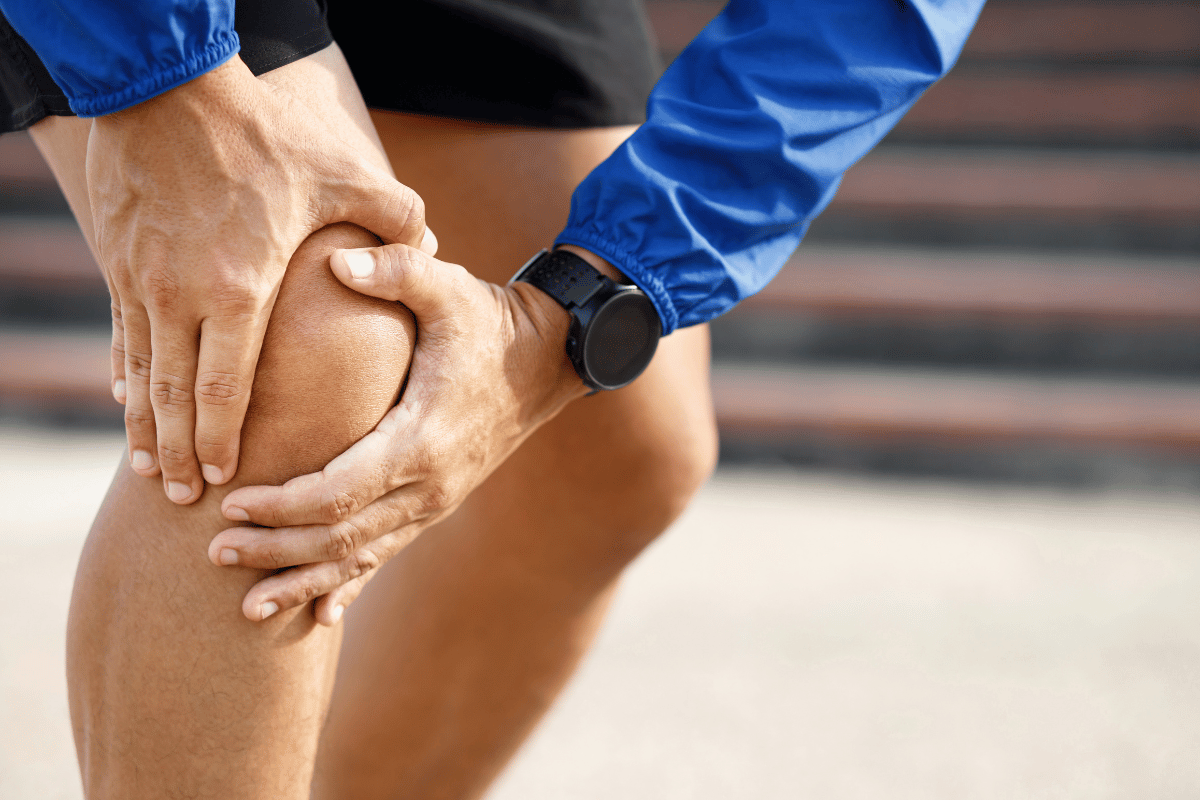 Man clutching his knee in pain, wearing a blue jacket and smartwatch, outdoors.