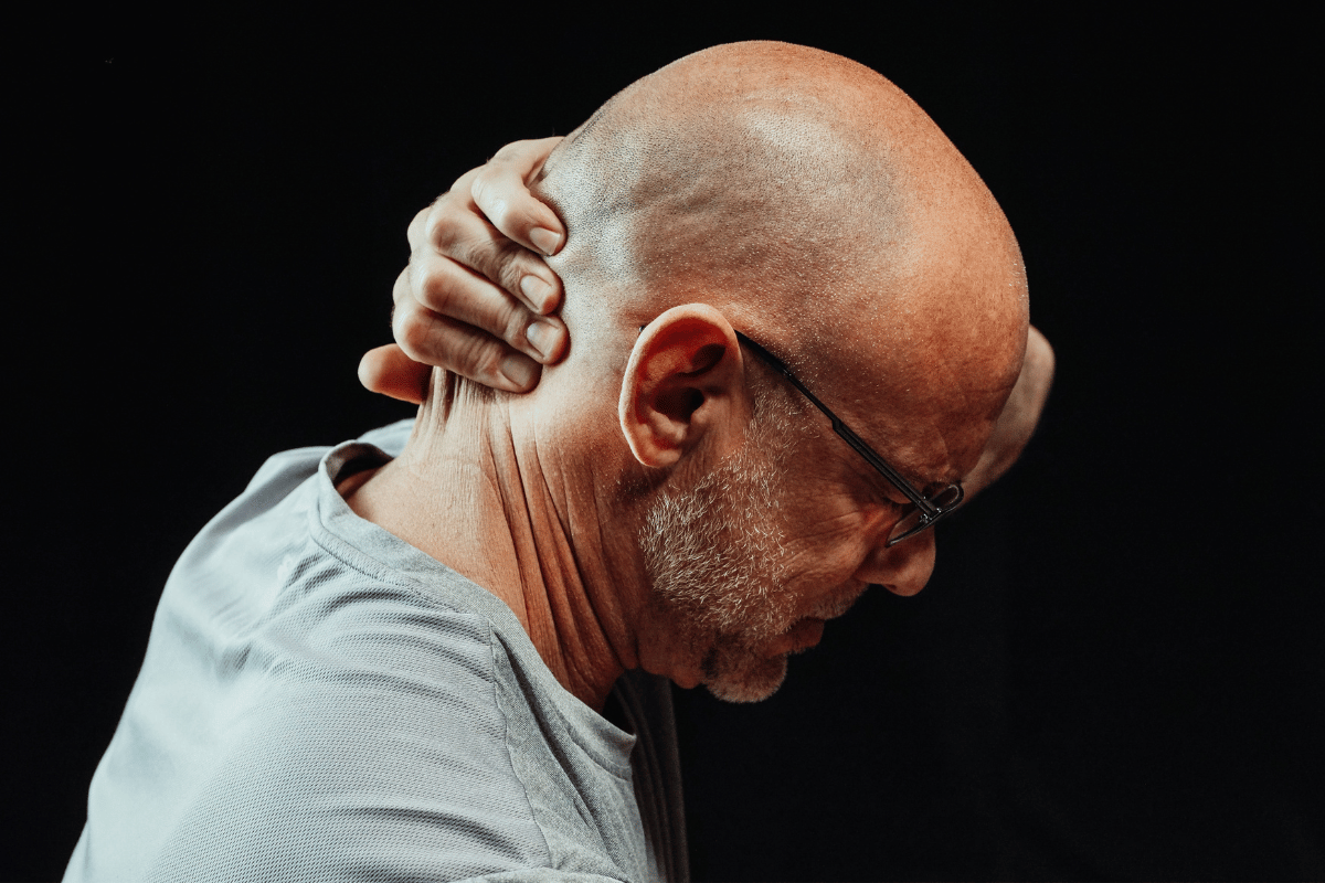 Bald man with glasses holds his neck in pain, against a black background.
