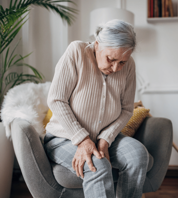 Elderly woman sitting, holding knee in pain, possibly indoors.