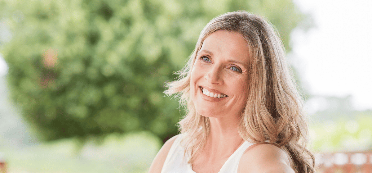 Woman with blonde hair smiles outdoors, green background.