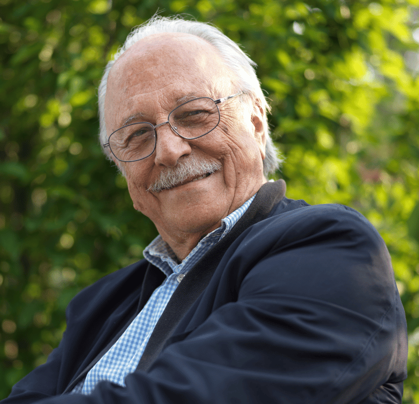 Smiling older man with glasses, white hair, and mustache, outdoors in a blue jacket.