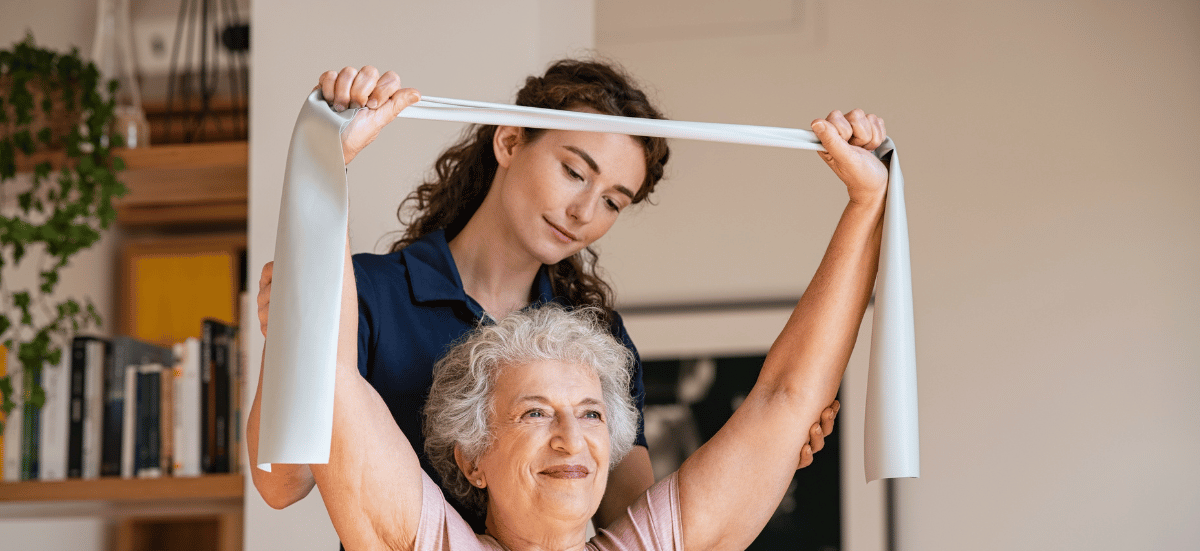 Woman assisting an elderly woman with arm exercises, using a white resistance band. Indoor setting.
