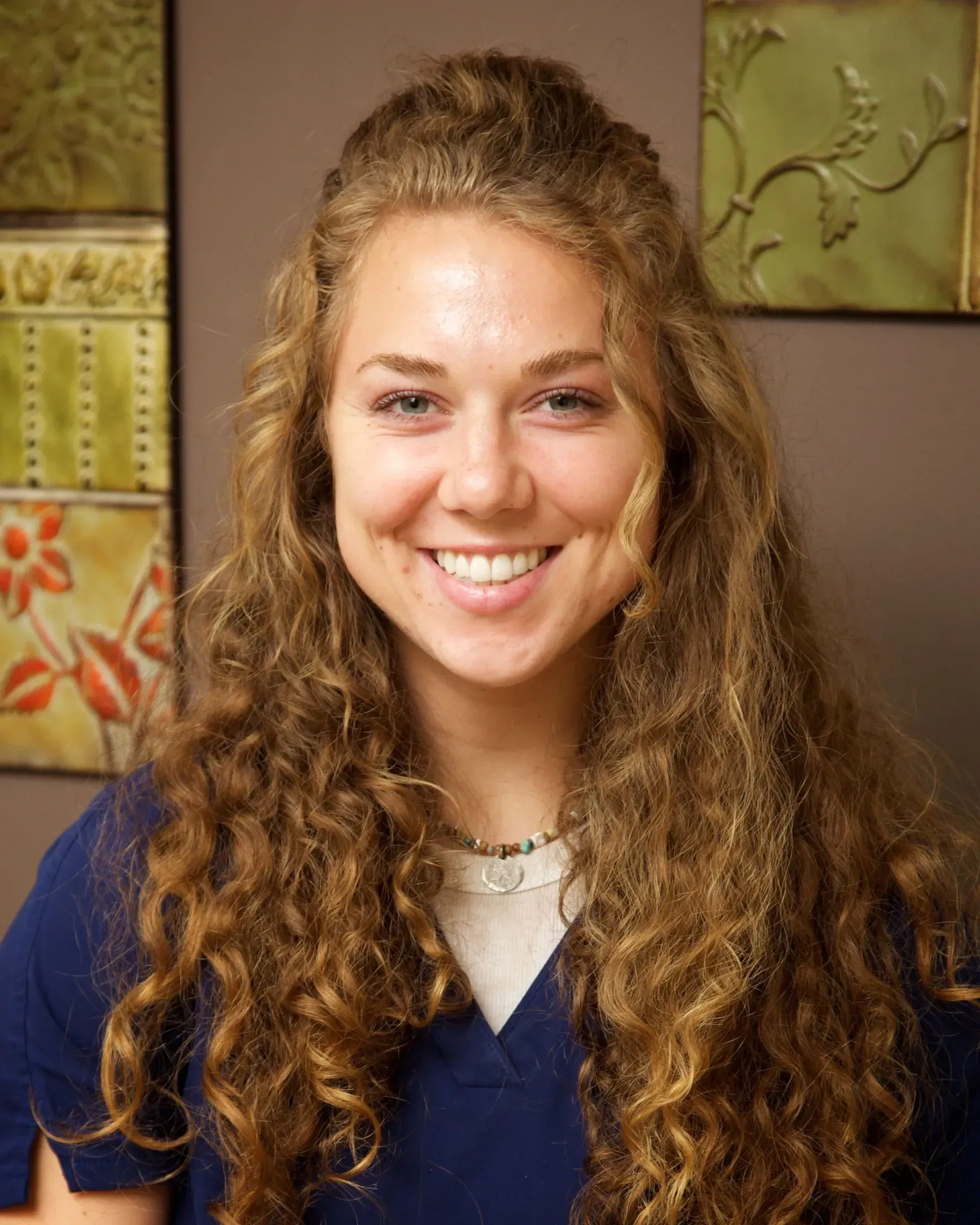 Woman with curly brown hair smiles at the camera, wearing a dark blue scrub top.