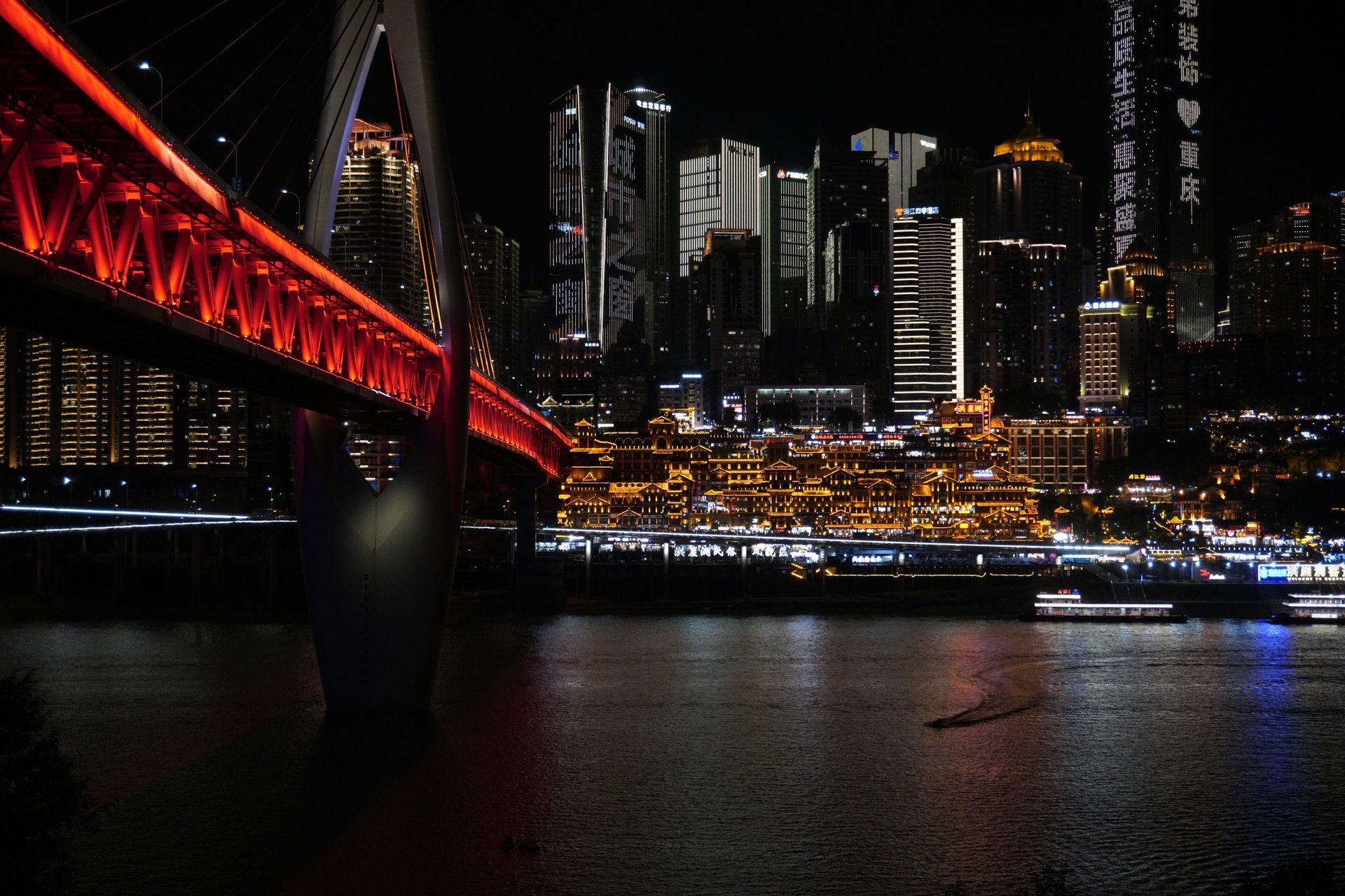Red-lit bridge over dark water at night, with illuminated city skyline in background.