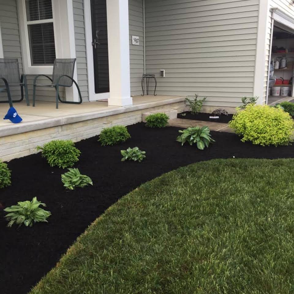 A front yard with a landscaped garden bed filled with plants and dark mulch, bordering a green lawn.
