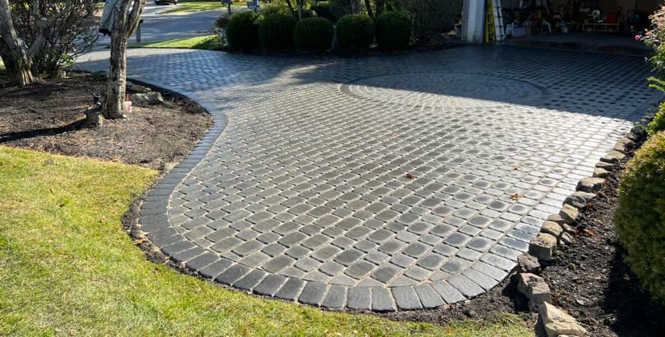 Brick driveway with curved black border, surrounded by grass and shrubs.