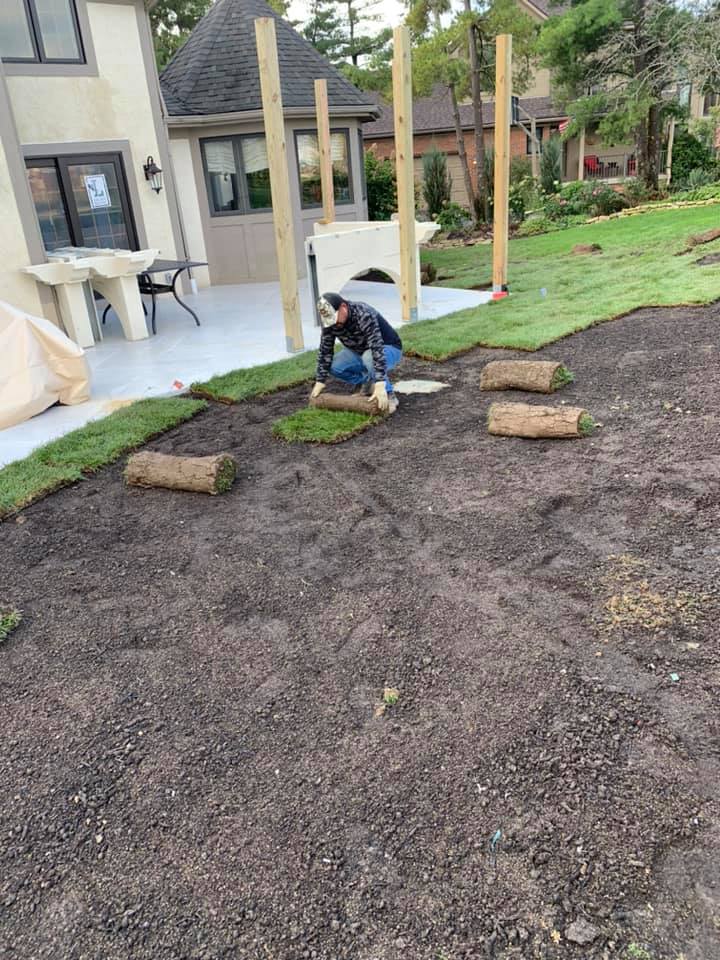 Person laying sod rolls in a yard near a patio and wooden posts.