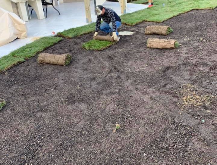 A person laying sod rolls on prepared soil, next to a patio and other rolls.