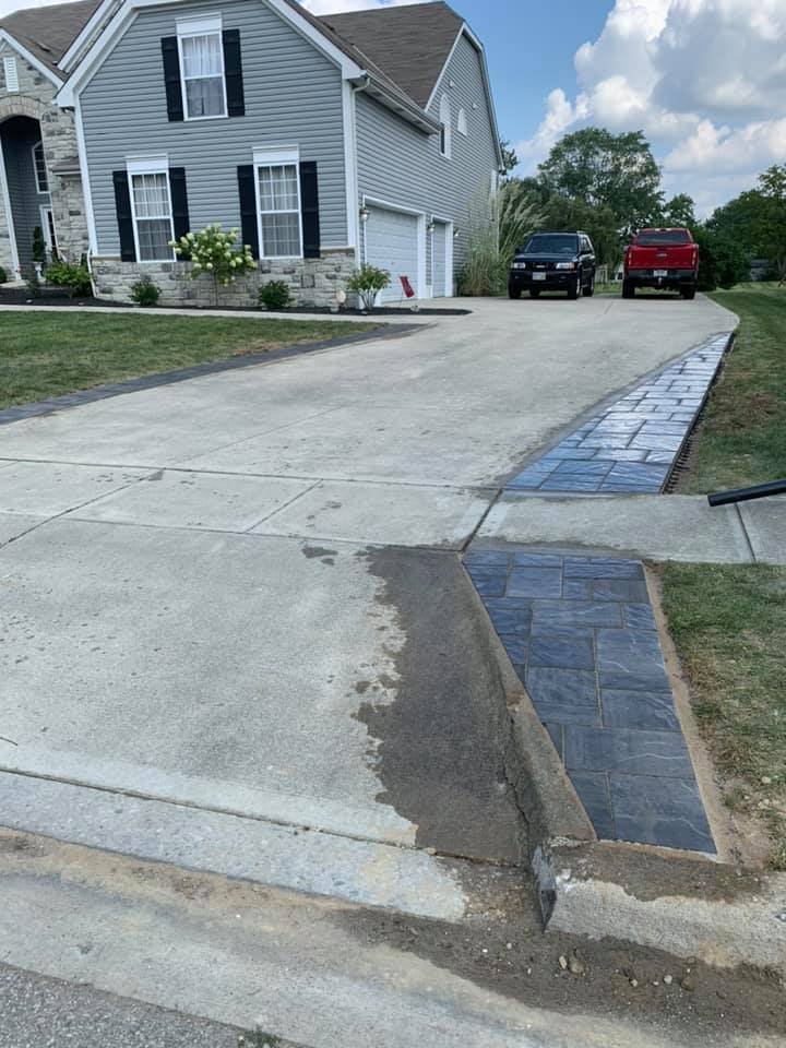 Driveway with a concrete surface and a brick border next to a house with parked cars.