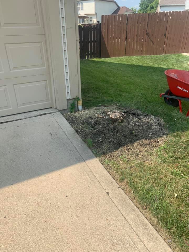 A concrete driveway borders a grassy yard with dirt pile next to garage and a wheelbarrow. Wooden fence in background.
