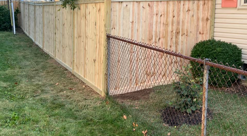 Wooden fence beside a chain-link fence in a grassy yard, near a house with bushes.