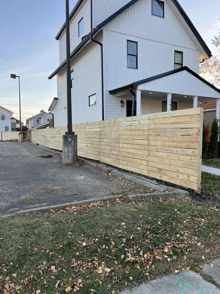 Wooden fence along the edge of a parking lot next to a white house with black trim.