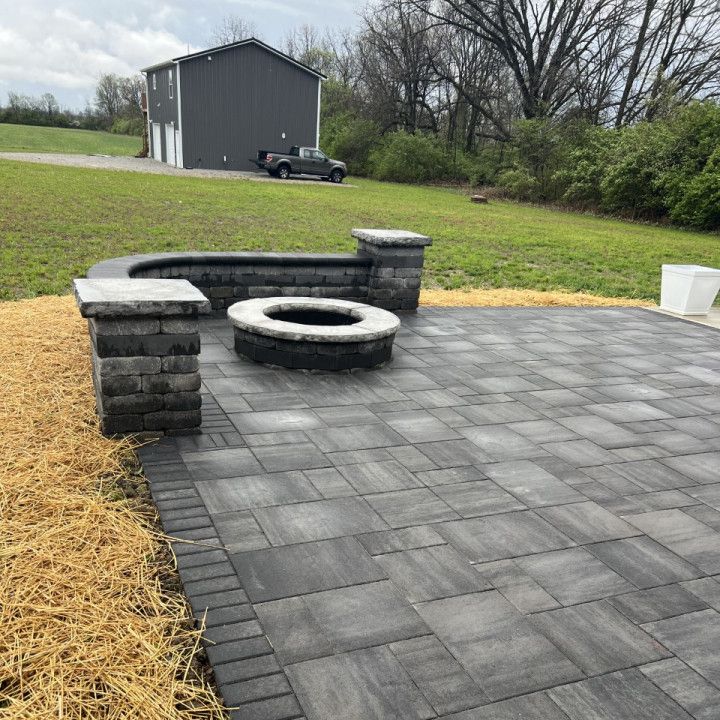 Patio with fire pit, seating, and gray pavers; building and truck in background.