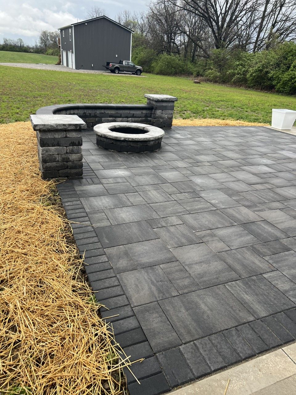 Patio with gray pavers, fire pit, and retaining wall; rural setting with a building in the background.