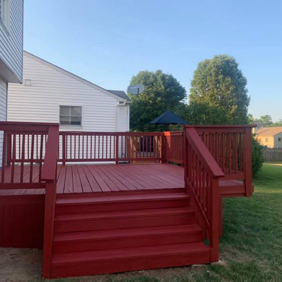 Red painted wooden deck with steps attached to a white house, surrounded by grass and trees.