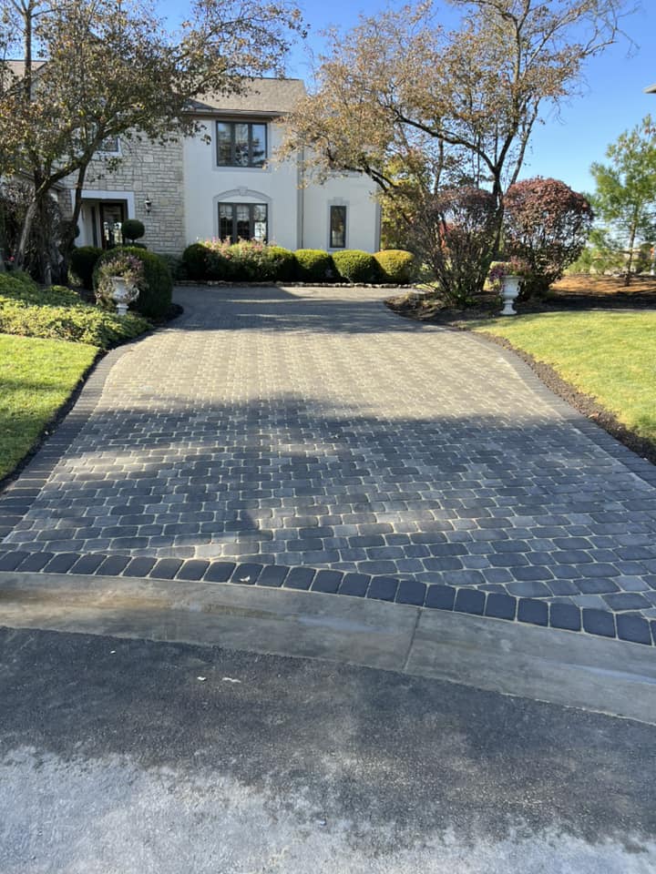 Brick driveway leading to a two-story house with green lawn and trees under a clear sky.