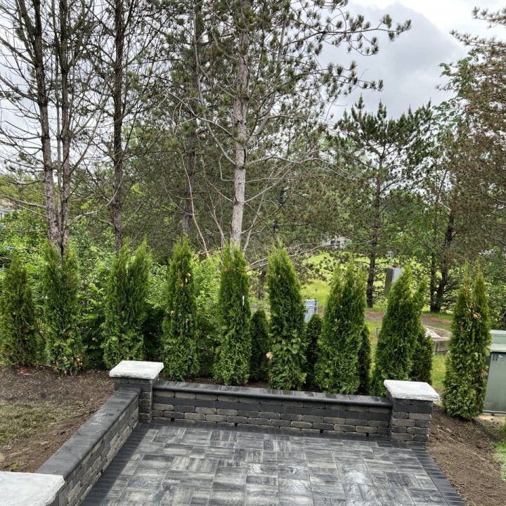 Stone patio with retaining wall, evergreen shrubs, and trees in the background under a cloudy sky.