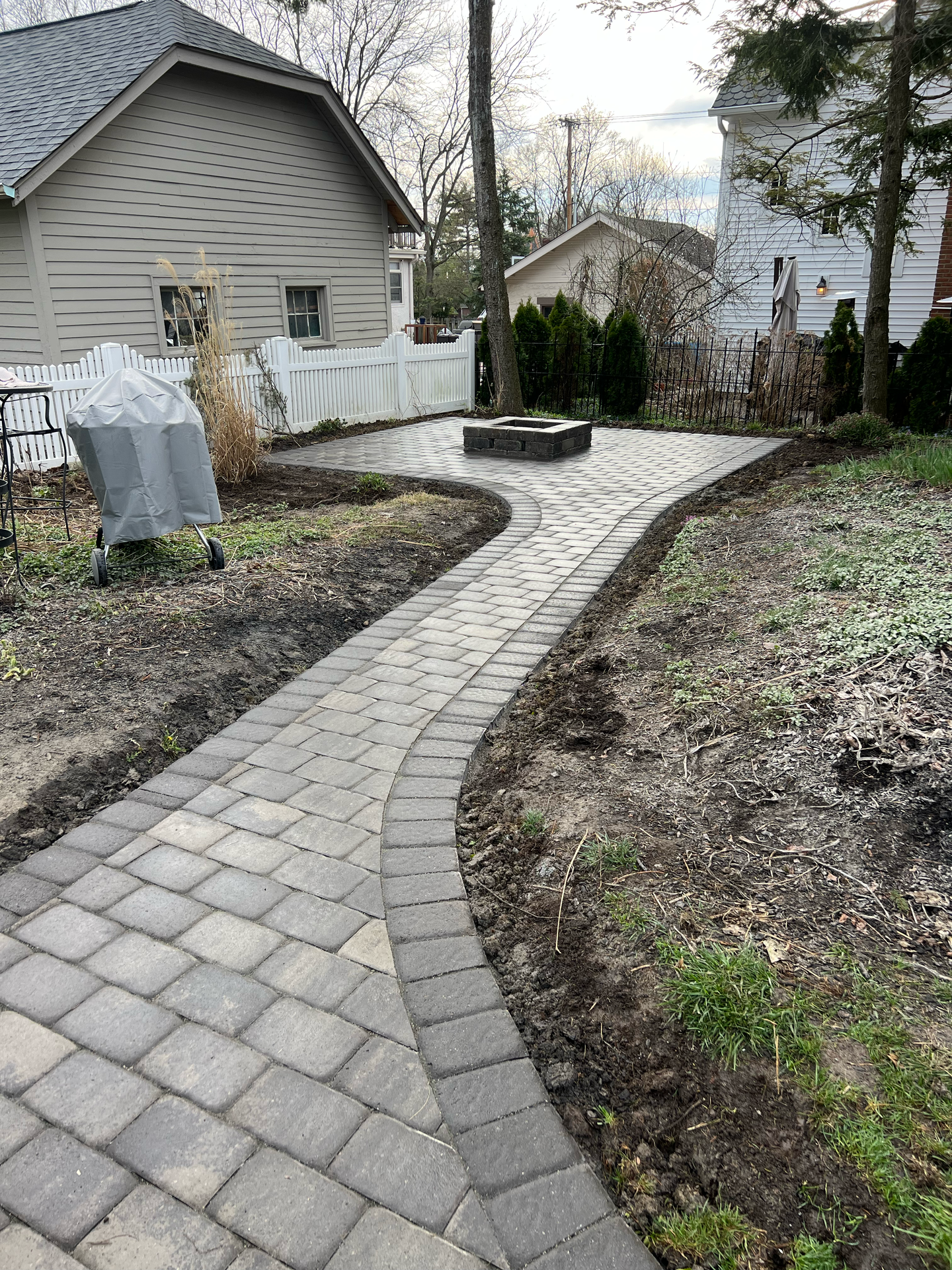 Brick pathway curves to a fire pit in a backyard, with houses and trees visible.