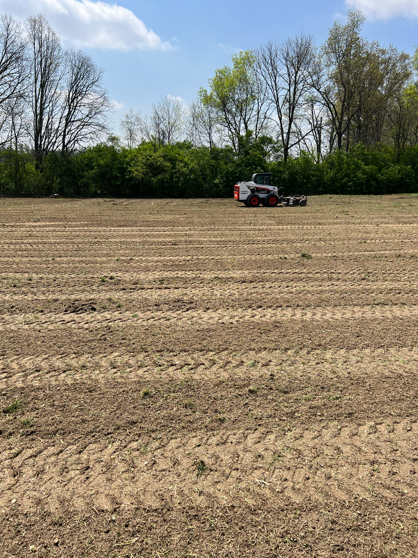 A small tractor working in a tilled field, trees and blue sky in background.