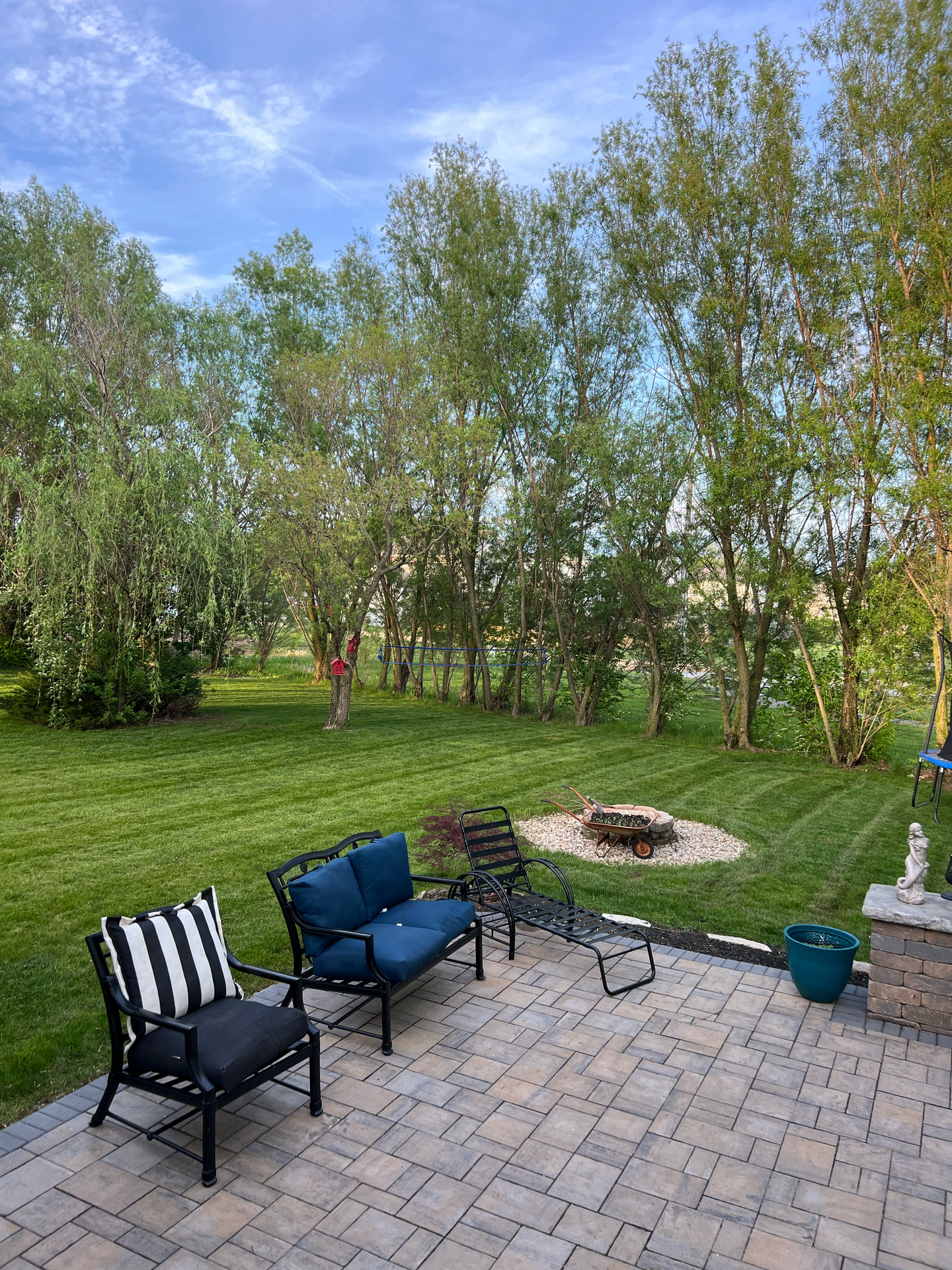 Patio with seating, fire pit, and trees in a backyard with green grass under a blue sky.