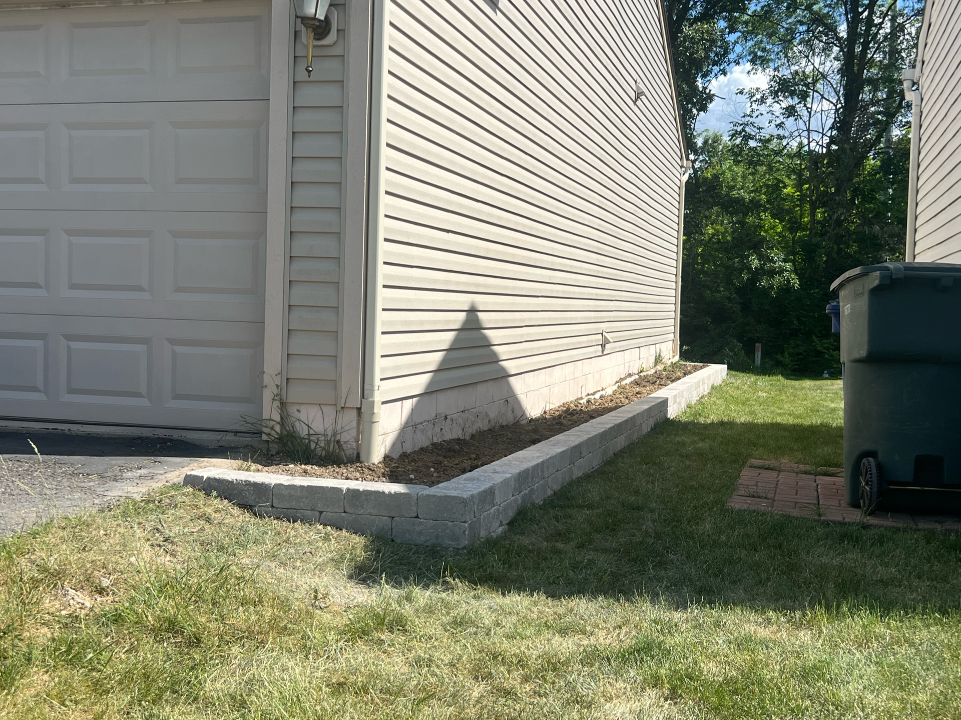 Gray retaining wall beside a tan building and garage door, with green grass and a trash bin.
