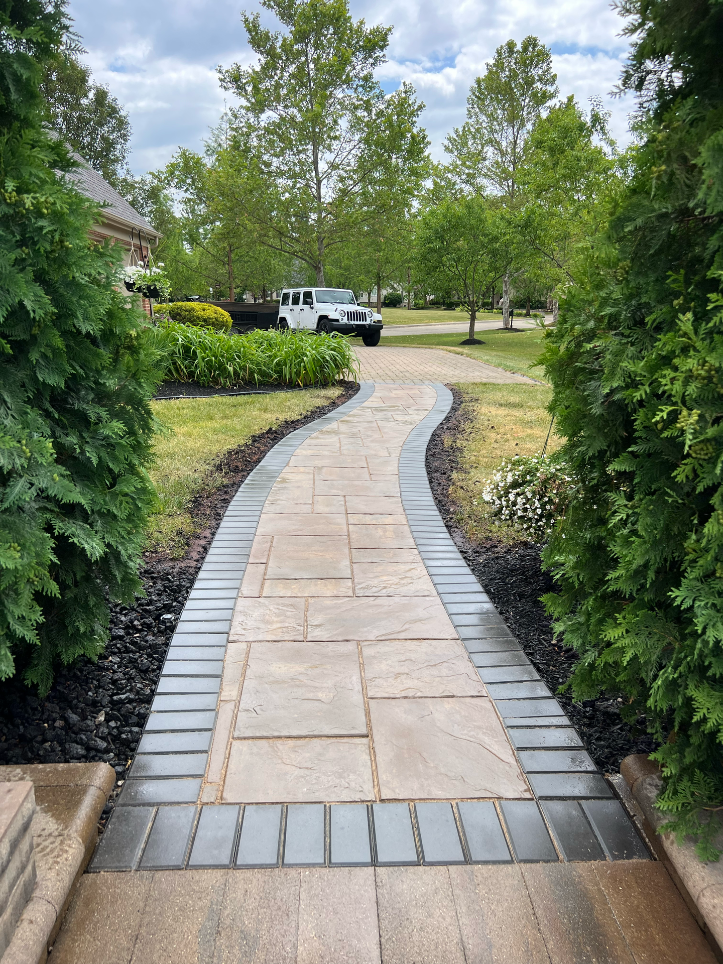 Stone pathway bordered by dark pavers and bushes, leading towards a white truck and trees.
