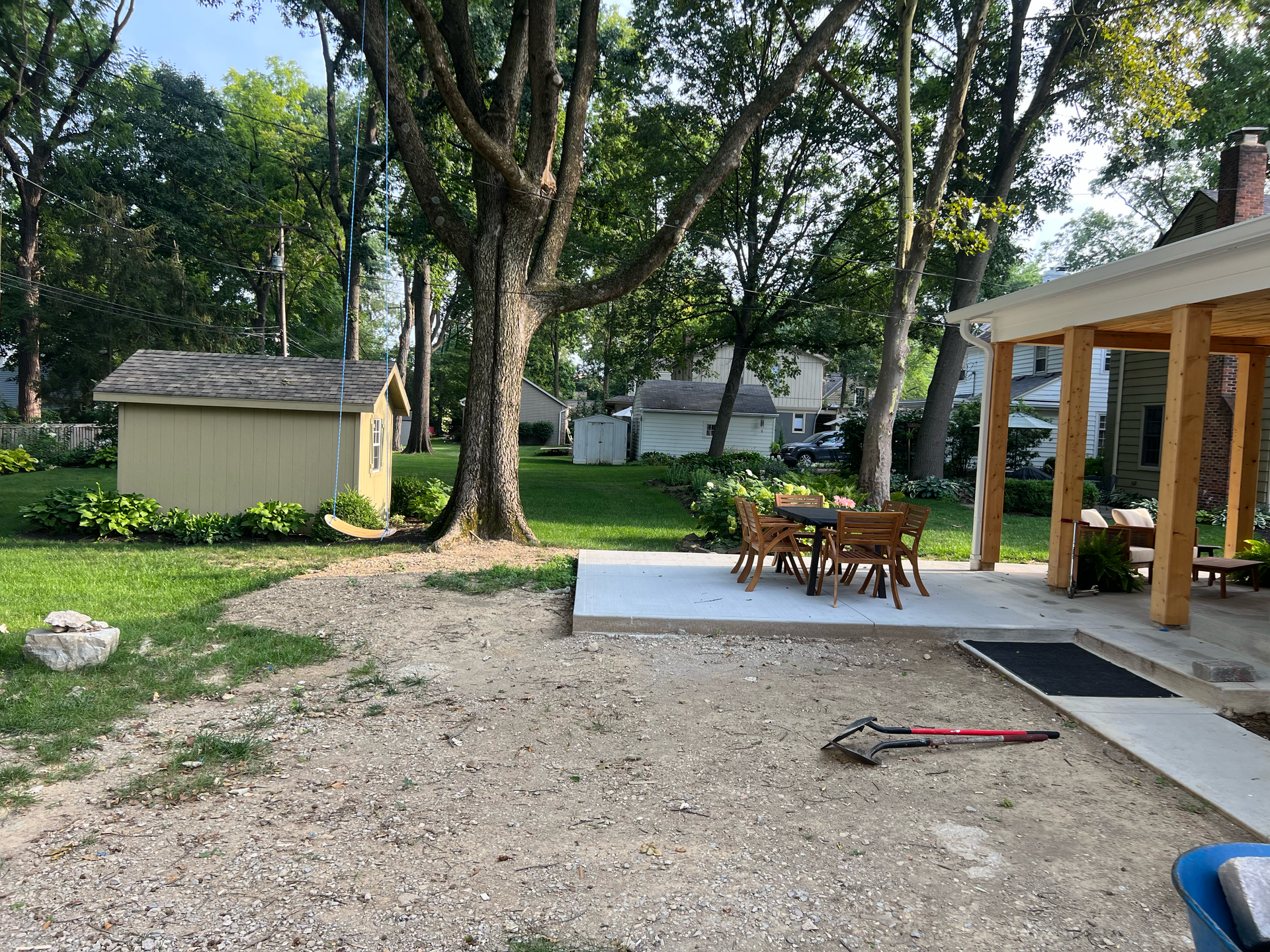 Backyard with a gravel area, concrete patio with table and chairs, shed, trees, and covered porch.