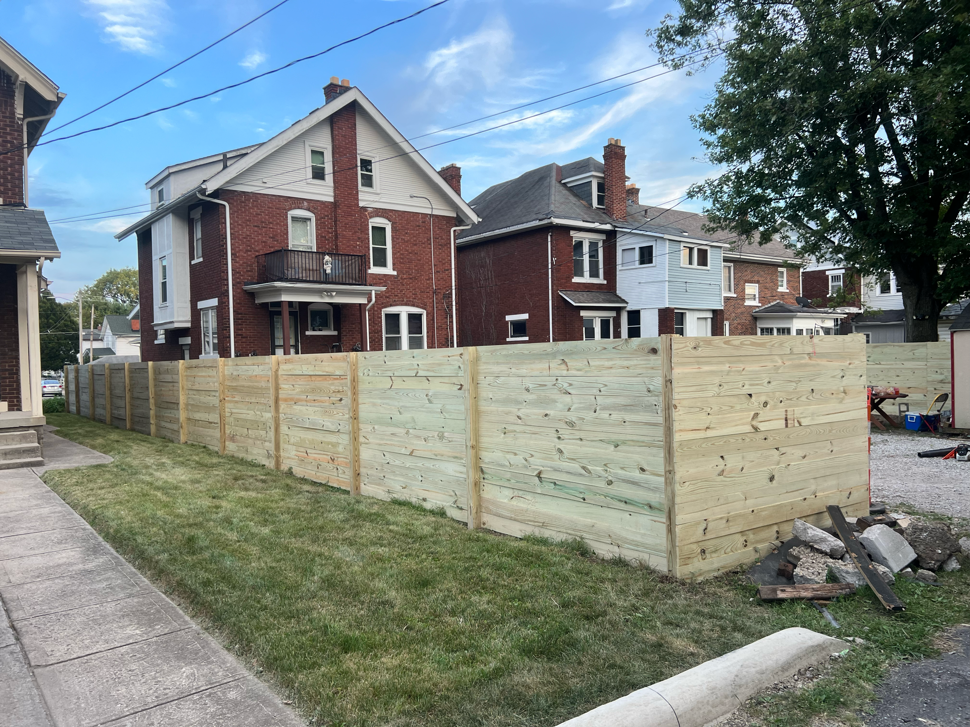 Wooden fence bordering a grassy lot in front of brick houses under a blue sky.