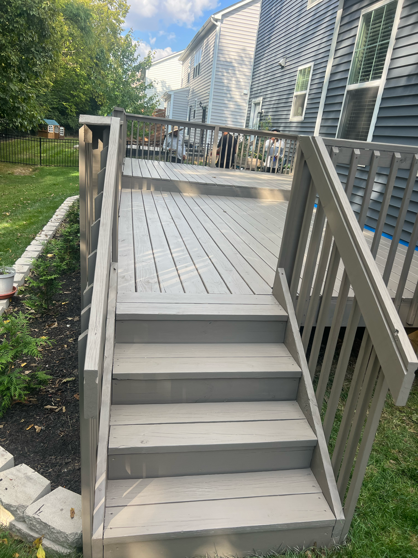 Gray wooden deck with stairs leading to a higher level; backyard setting with houses in the background.