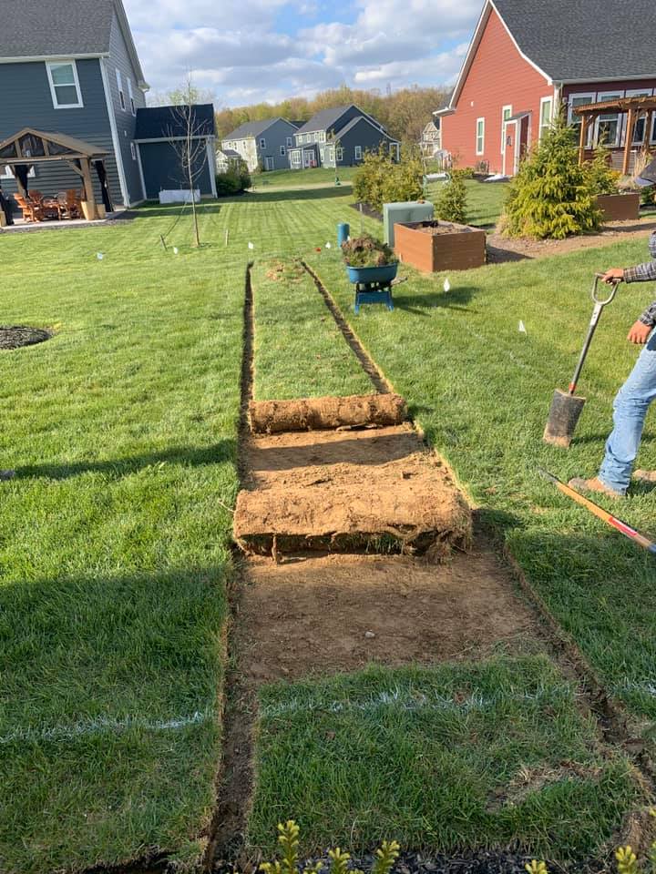 A person digs in a backyard, creating a trench with sod removed. Green grass surrounds.