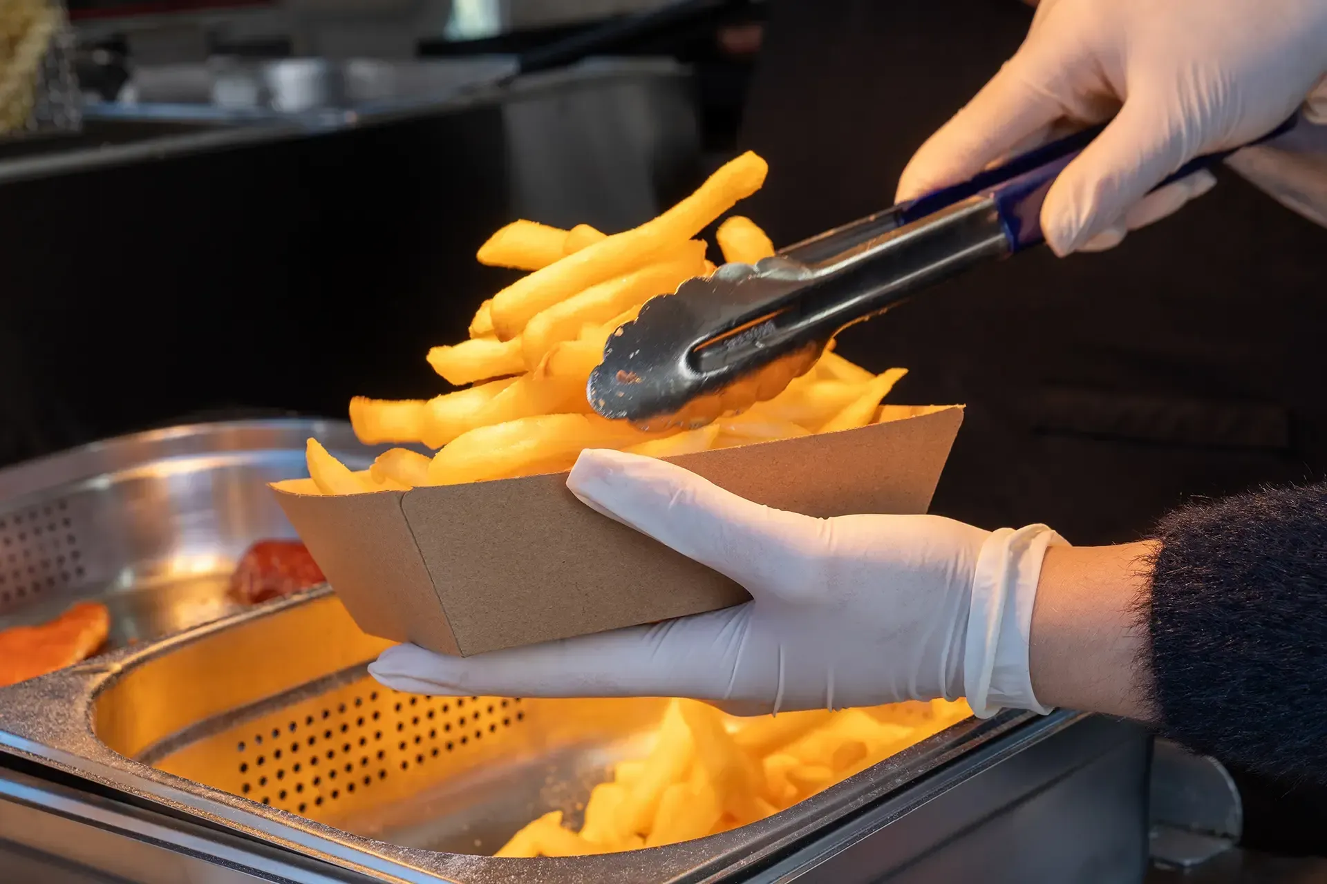 A person wearing gloves is holding tongs over a tray of french fries.