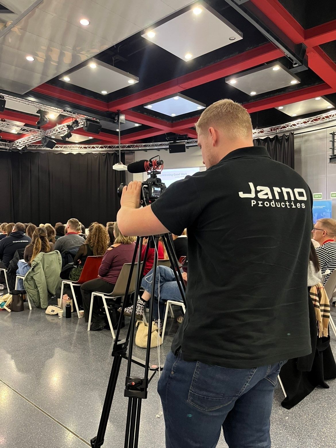 Iemand filmt een presentatie met een statief. Publiek op de achtergrond. 