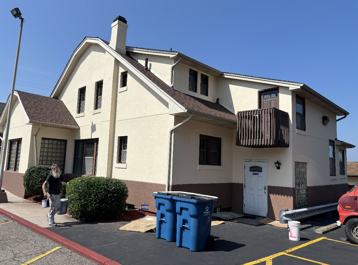 Two-story building with tan stucco and brown trim. Blue bins and a person are visible on the side.