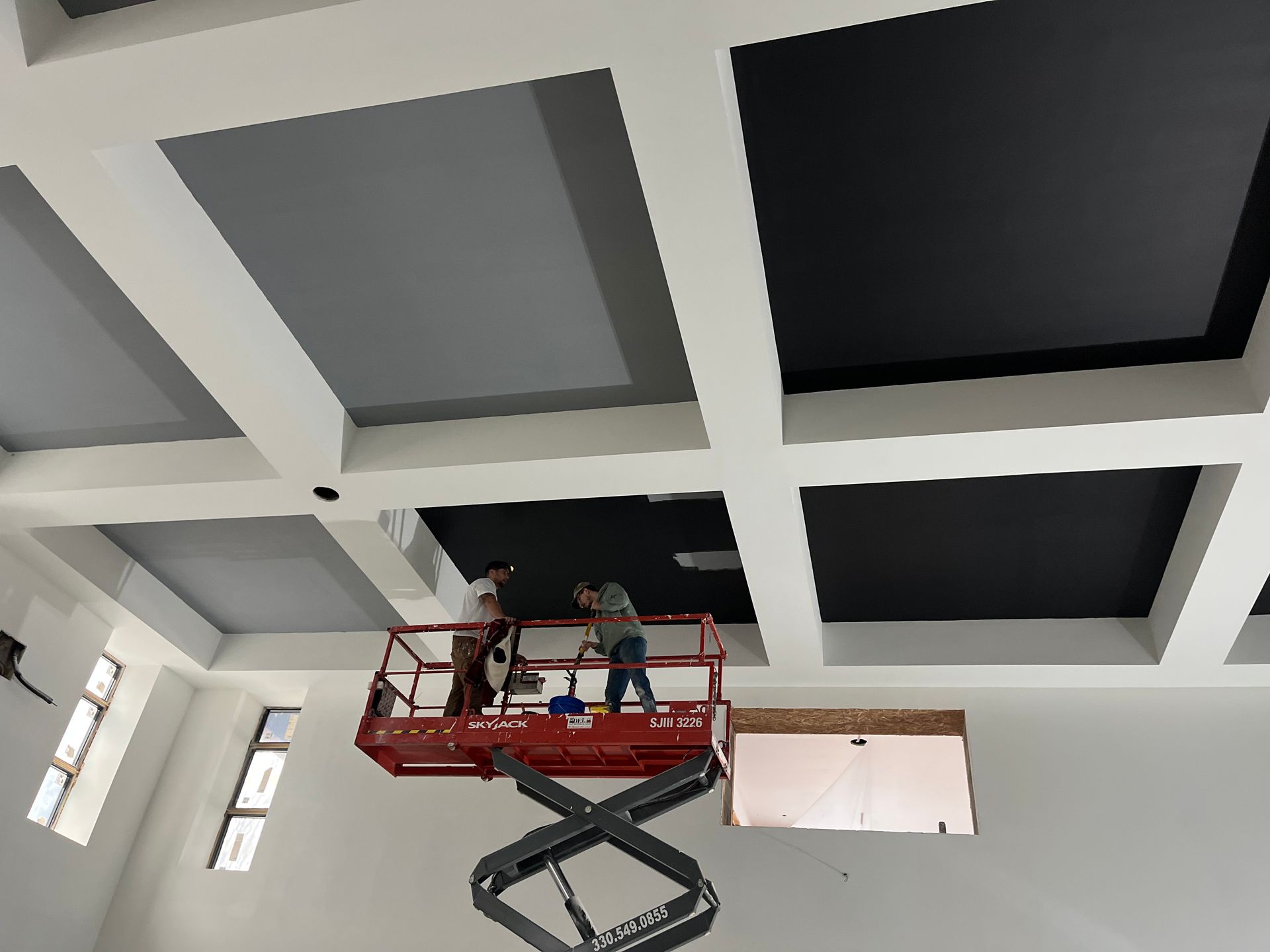 Two people on a lift, painting a ceiling with gray and black panels, white beams, interior.