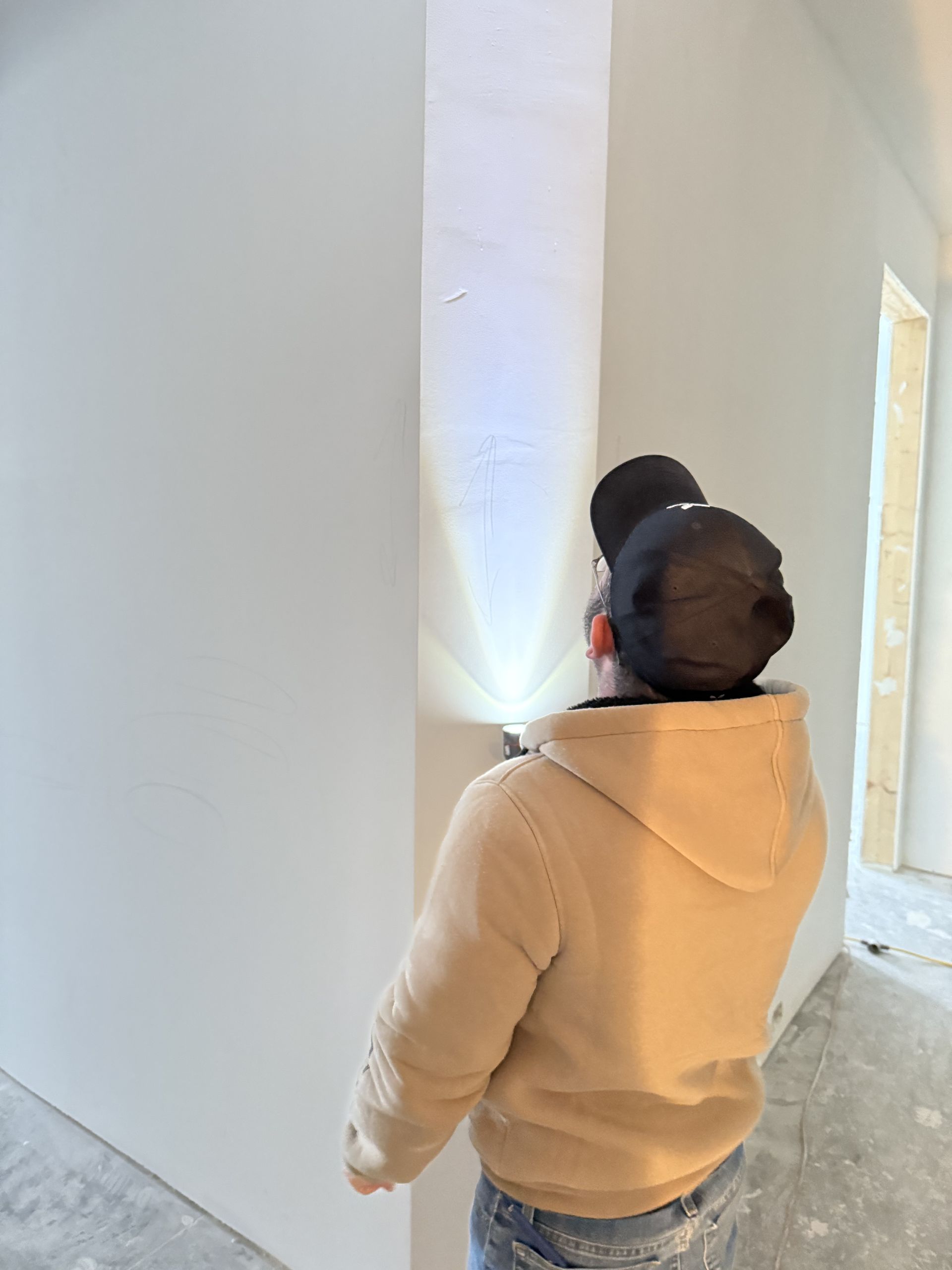 Person inspecting newly installed drywall with a flashlight. Construction setting, beige jacket.