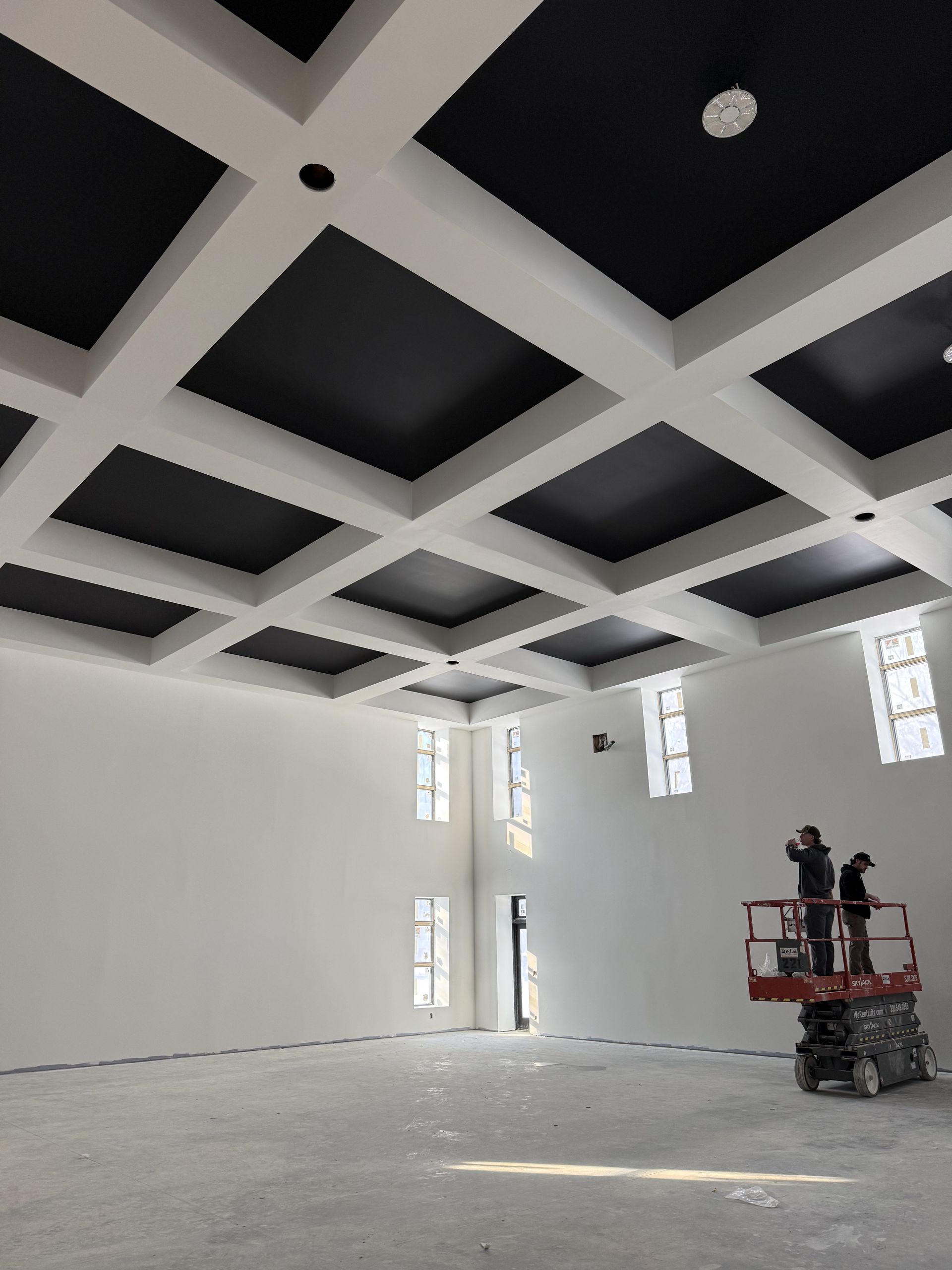 Room with white walls and a black coffered ceiling. Two people on a lift. Windows in the background.