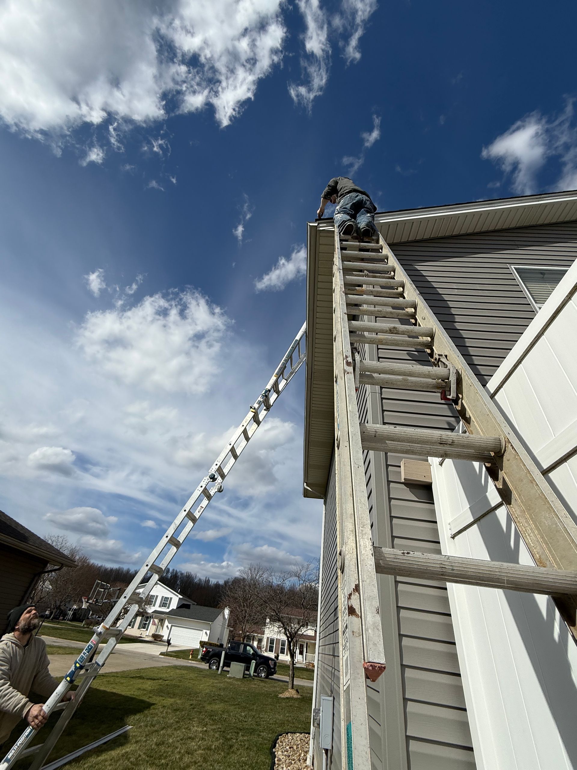 Person on ladder near a building's roof under a blue sky, other people visible on the ground.