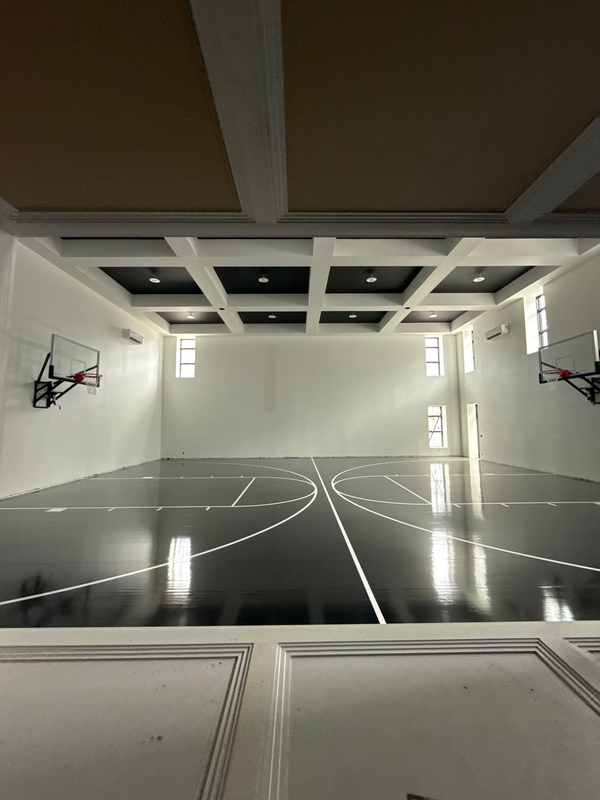 Indoor basketball court with black floor, white walls, and two hoops. Ceiling has recessed lighting.