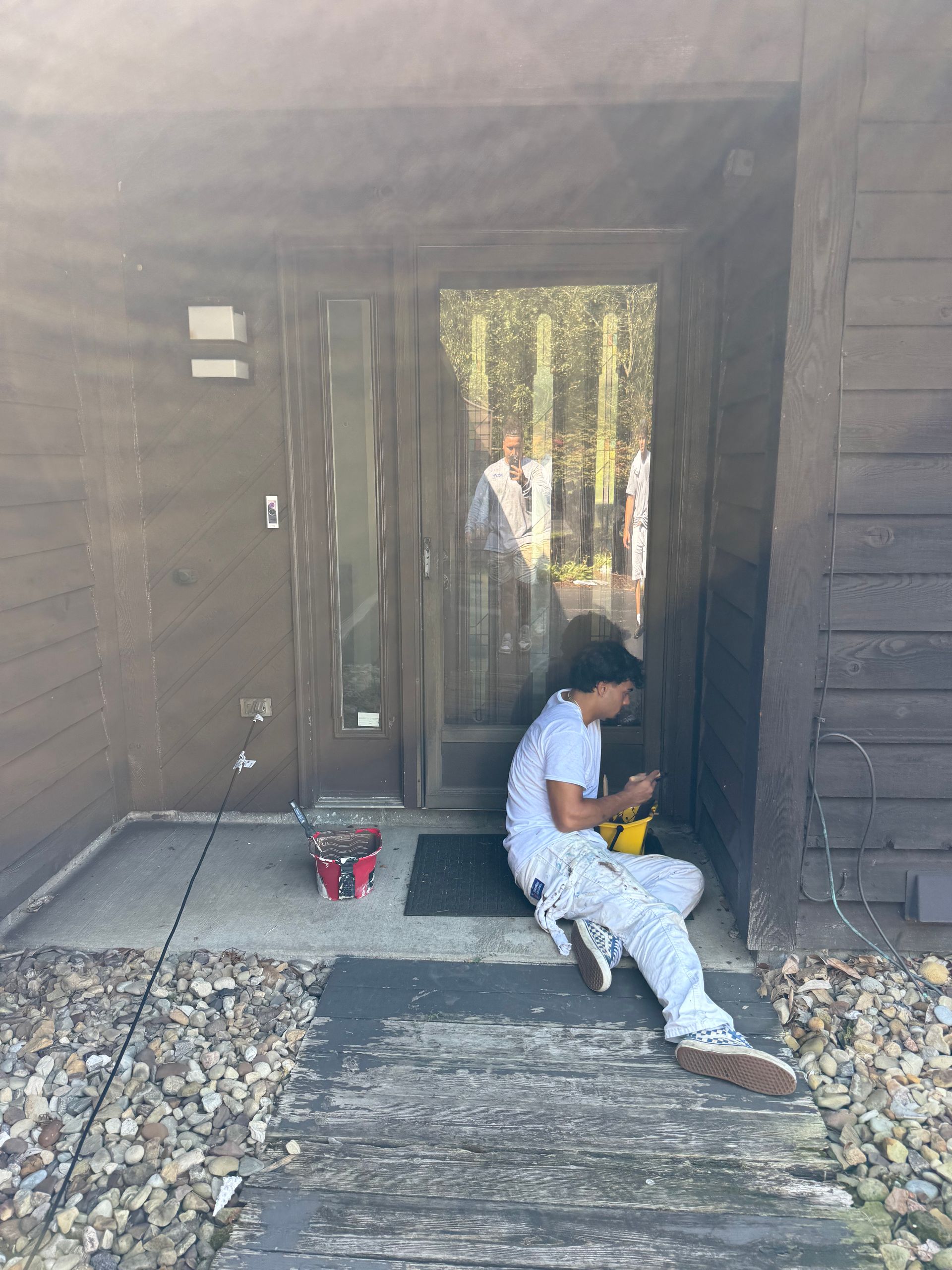 Person sits on a porch, working with a yellow tool. A person is reflected in the glass door.