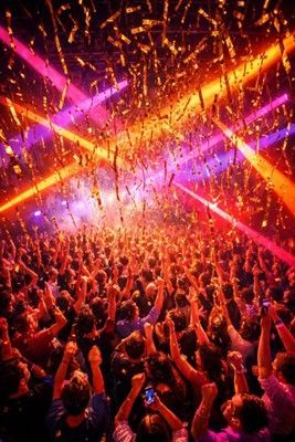 A crowd at a nightclub raises their hands as gold confetti falls under vibrant pink and orange laser lights.