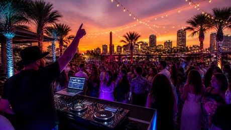 DJ performing at sunset beach party with crowd, palm trees, and city skyline in the background